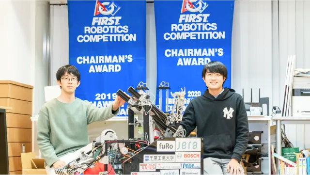 2人の学生が中央のロボットを挟んで座っている写真。